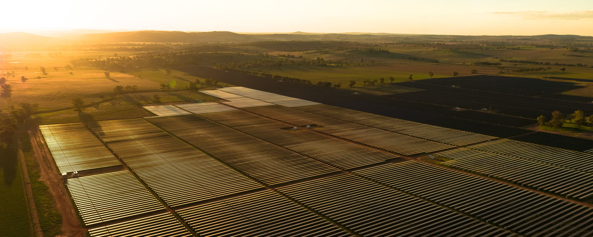 夕日を浴びた広大なソーラーファームの俯瞰図。整然と並んだソーラーパネルが平坦な土地に広がり、周囲には広い農地と遠くの丘陵が黄金色の空の下に見える。