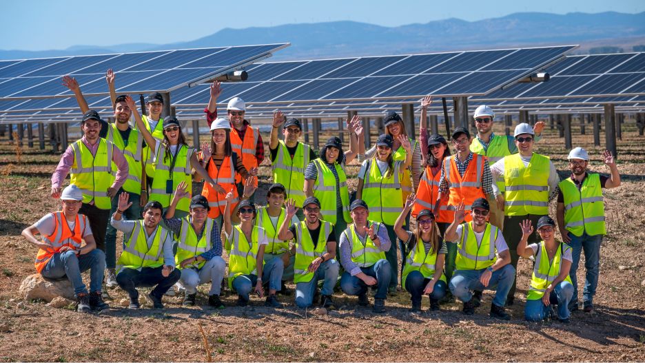 A group of people in safety vests and helmets stands cheerfully in front of solar panels in a sunny field.