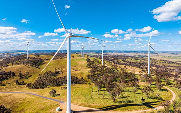 A wide aerial view of a wind farm with multiple large white wind turbines spread across rolling green hills under a blue sky with scattered white clouds.