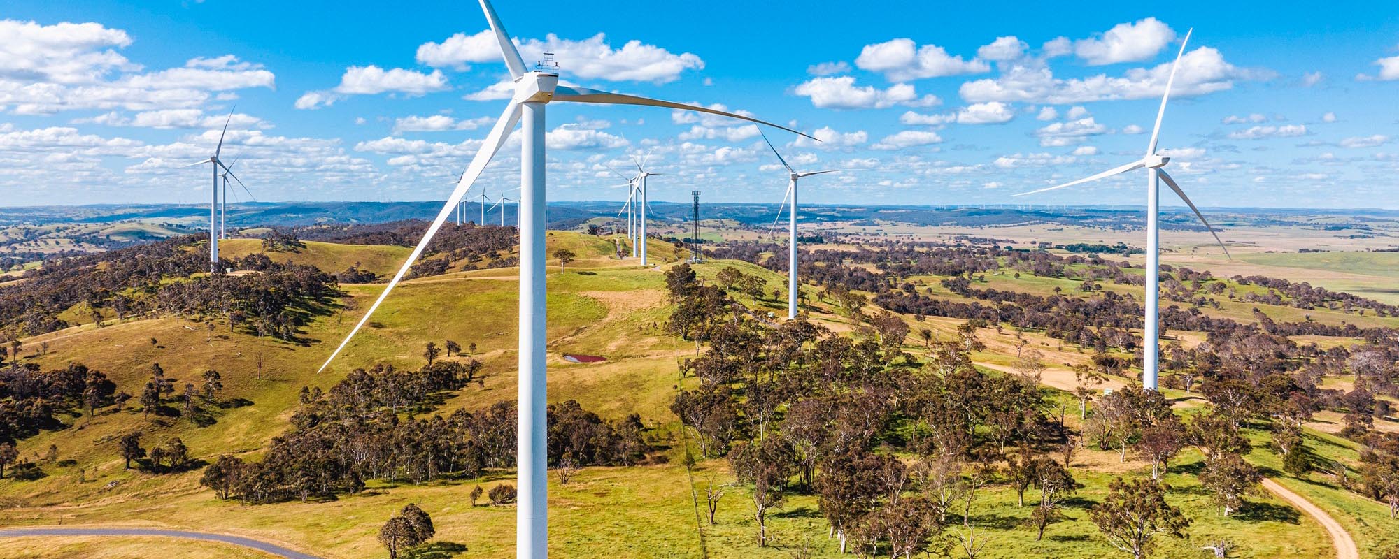 A wide aerial view of a wind farm with multiple large white wind turbines spread across rolling green hills under a blue sky with scattered white clouds.