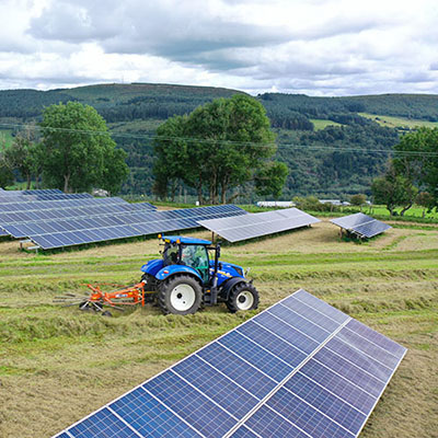 A blue tractor mowing grass in a solar farm surrounded by green hills and trees under a partly cloudy sky, demonstrating agricultural activity integrated with solar panel installations.