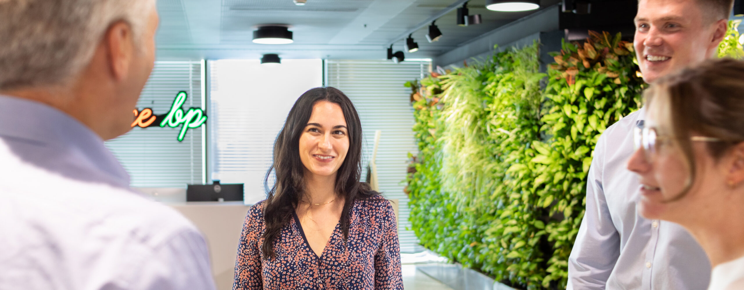 オフィスの中で会話をしている数人の人々。背景には緑豊かな植物の壁があり、自然を感じられる明るい空間が広がっている。