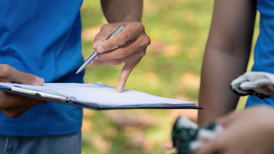 青いシャツを着た人が屋外でクリップボードを持ち、ペンで書類を指しているクローズアップ写真。