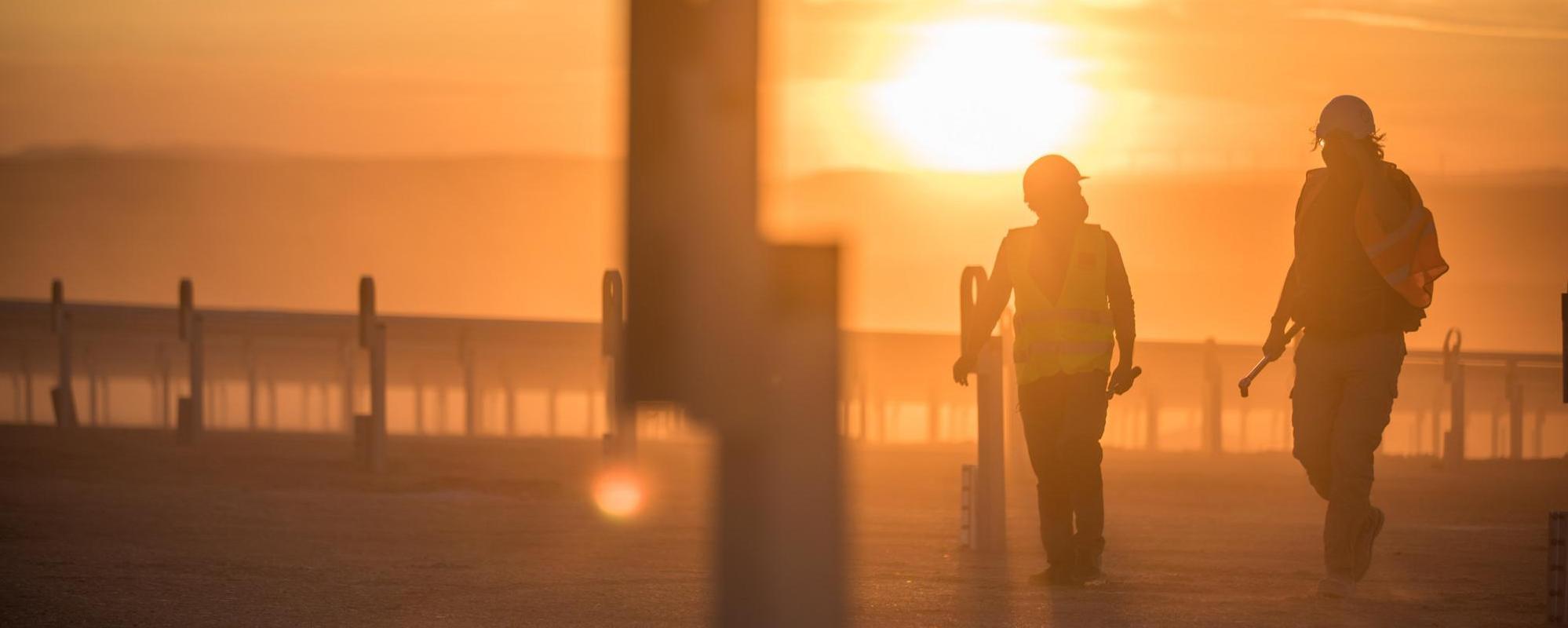 Two workers in safety gear walk through a construction site of a solar farm at sunset, silhouetted against an orange sky.